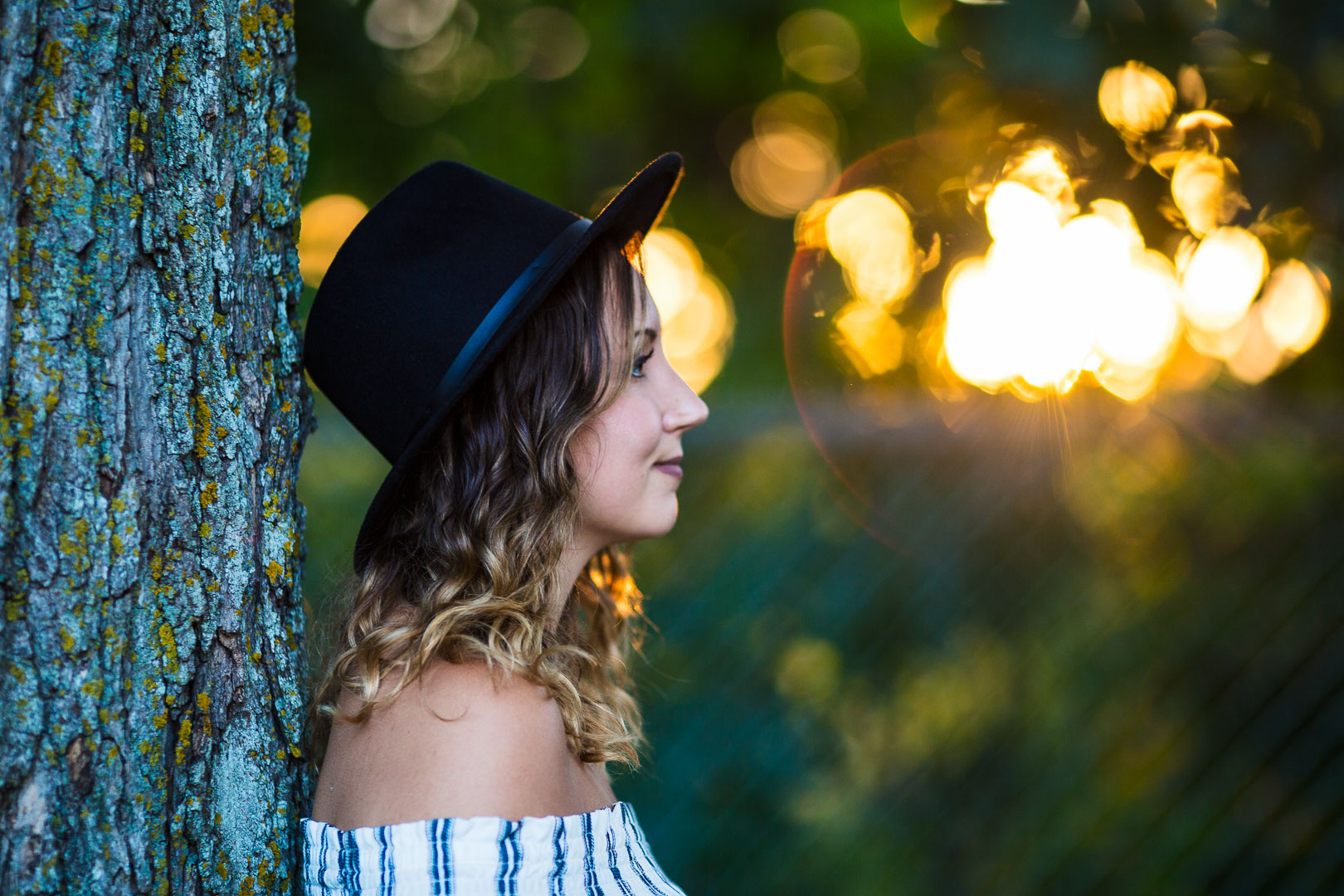 Portrait de profil d'une femme avec chapeau noir appuyée contre un arbre, soleil en contre-jour créant un bokeh doré, fill flash éclairant le visage