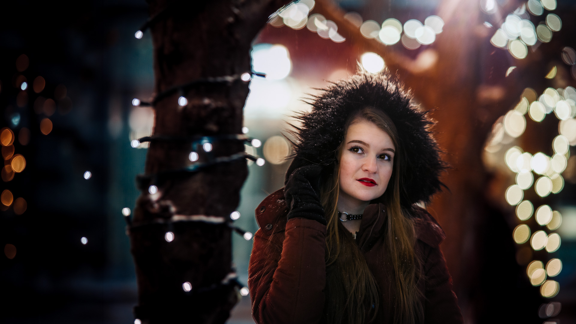 Portrait de femme en manteau d'hiver avec capuche de fourrure, éclairée au flash sur fond de lumières de Noël en bokeh, nuit hivernale