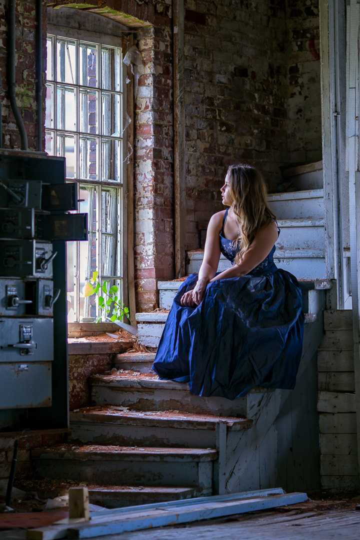 Femme en robe bleue assise près d'une fenêtre dans un bâtiment abandonné, éclairée par la lumière naturelle latérale